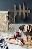 Jam and bread on wooden board on marble worksurface below magnetic knife rack on blue-grey wall