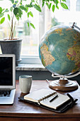 Globe next to laptop and pocket diary on desk below window