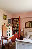 Old dolls' house on console table, red bookcase and white wooden bed in bedroom