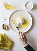 Hand using fork to take piece of yummy fresh burrata from plate near bread and oil against white background