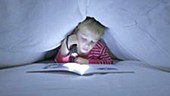 Boy reading book under sheet with torch