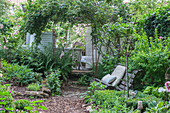 Antique garden bench in front of rose arch and white wooden pavilion with seating area, ferns, perennials and box hedge