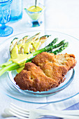Breaded chicken fillet with green asparagus and grilled avocado on a glass plate