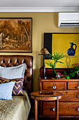 Hunting paintings over a leather bed next to a chest of drawers and wooden side tables