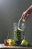 Ingredients for green spinach kale apple honey smoothie in glass blender, young man pouring milk