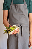 Man in grey apron before white background holding a bundle of green asparagus wrapped in a towel in his hands