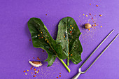 Fresh spinach, spices and a meat fork on a violet surface