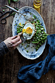 From above person hands eating bread toast with sauteed green peas and fried egg on wooden table
