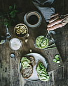 From above hand of unrecognizable person cook making toasts with green cashew pate and slices of cucumber on wooden board