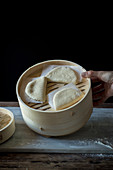 Crop person cook holding bamboo steamer with raw lotus leaf buns for Gua Bao at table