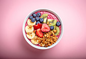 Summer acai smoothie bowl with strawberries, banana, blueberries, kiwi fruit and granola on pastel pink background