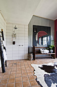 Wooden bench, round mirror, corrugated metal wall, cowhide rug and tiled floor in spacious bathroom