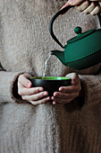 Green black cup in hands of woman in sweater and teapot with pouring water