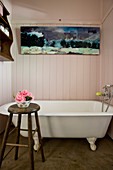 Vintage bathtub below painting in wood-clad bathroom with bowl of flowers on stool