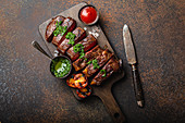Grilled or fried and sliced marbled meat steak with fork, tomatoes as a side dish and different sauces on wooden cutting board, top view, close-up, stone rustic background