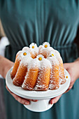 A woman holding a lemon Bundt cake decorated with daisies for a birthday
