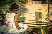 Girl in white summer dress sitting on jetty next to idyllic pond