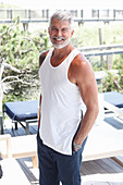 A grey-haired man on a terrace wearing a white vest and jeans