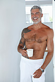 A topless, grey-haired man wearing white trousers and holding a cup of coffee