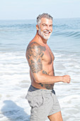 Grauhaariger Mann mit großem Tattoo in hellgrauen Shorts am Meer