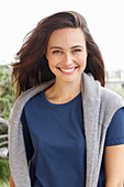 A young brunette woman wearing a short-sleeved blue t-shirt with a grey jumper around her shoulders