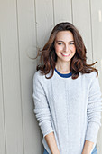 A young brunette woman standing in front of a wooden wall wearing a light knitted jumper and jeans