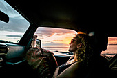 Frau sitzt im Auto beim Sonnenuntergang, Fuß ragt aus dem Fenster