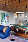 Blaues Sofa und Beistelltisch, dahinter Esstisch mit Stühlen in offenem Wohnraum mit Holzdecke und Wänden in Beton-Optik