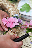 Strohkranz wird mit Hortensien, Vergissmeinnicht und Schnittlauchblüten gebunden