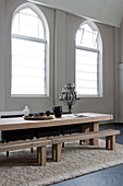 Long wooden table and benches on rug next to arched windows