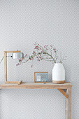 Table lamp and vase with a sprig of rosehips on a console table in front of patterned wallpaper