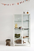 White display case with vintage crockery and rustic stool under a pennant chain