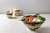 Poke bowl with soy sauce marinated salmon, rice, avocado and tofu cheese served in ceramic bowl with chopsticks and teapot on white marble table
