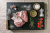 Raw uncooked rack of lamb on black burned wooden cutting board with salt, herbs rosemary, pepper and garlic