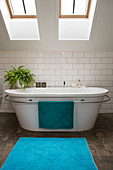 A bathtub with a towel rail and an indoor fern under skylights in a bathroom with white underground-style tiles