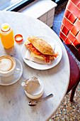 Breakfast consisting of a sandwich, coffee, orange juice and yoghurt on a bistro table