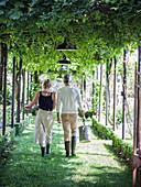 Zwei Personen mit Gartenutensilien laufen im Laubengang unter Pergola
