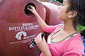 Teenage girl putting brown bottles into a recycling bank