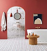 Frei stehende Badewanne und Rattantisch im Bad mit ziegelroter Wand und Marmorfliesen, Blick durch Rundbogenöffnung auf Waschbecken