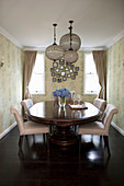 Antique, oval dining table with pale upholstered chairs and pendant lamps in dining room with patterned wallpaper