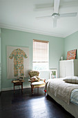 Bett, weißer Schrank, Polsterstuhl vor Fenster und Kinderkleidung als Wanddekkoration in pastellgrünem Zimmer