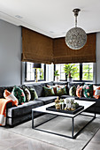Colourful scatter cushions on grey velvet sofa in front of corner windows with fabric blinds