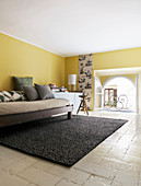 Sofa bed against mustard-yellow wall in attic room with stone floor and arched window