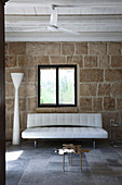 White, modern sofa and designer standard lamp against rustic stone wall