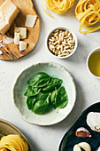 Top view of fresh green basil leaves on plate next to pine nuts and Parmesan cheese on white kitchen table