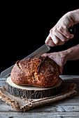 Unrecognizable person using knife to cut loaf of fresh sourdough bread on piece of wood against black background