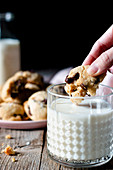 Unrecognizable female dipping piece of yummy vegan cookie with chocolate chips into glass of fresh milk on wooden table against black background