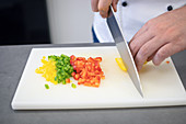 Crop cook in uniform grinding red green yellow peppers with large knife on white board