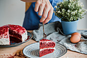 Unrecognizable person putting plate with piece of delicious red velvet cake on table in kitchen