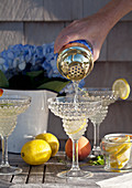 Vodka lemonade being poured from a shaker into a glass on an outdoor table with flowers, lemons, peaches, thyme and mint